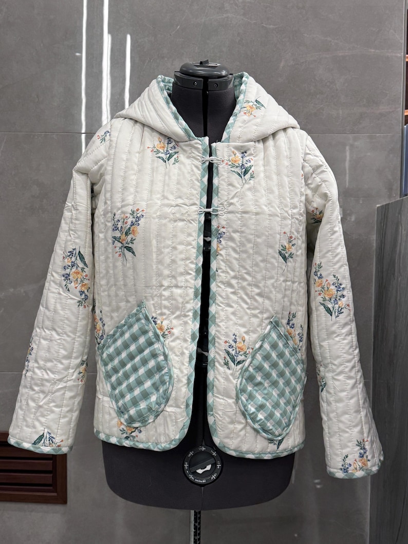 K&ouml;nnte beinhalten: Eine wei&szlig;e gesteppte Jacke mit Blumendruck und gr&uuml;nen Karotassen. Die Jacke hat eine Kapuze und einen Knopfverschluss.