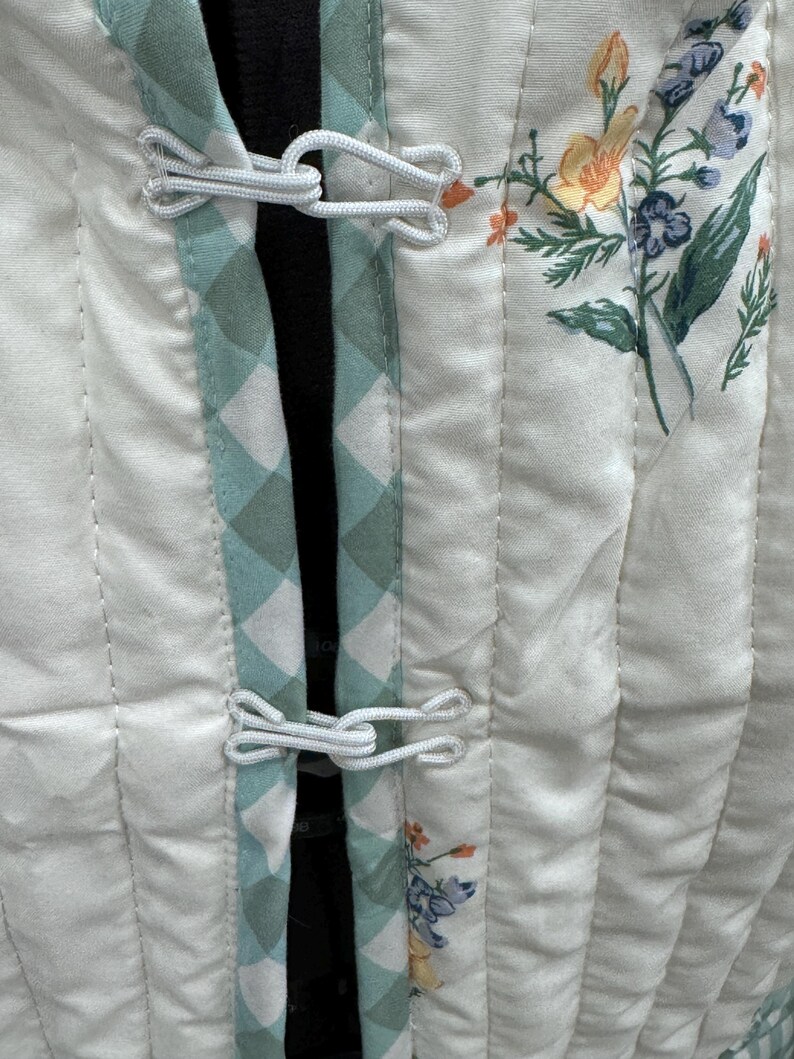 K&ouml;nnte beinhalten: Eine wei&szlig;e gesteppte Jacke mit gr&uuml;nem und wei&szlig;em Karomuster am Saum. Die Jacke hat einen Blumendruck und wei&szlig;e Knopfverschl&uuml;sse.