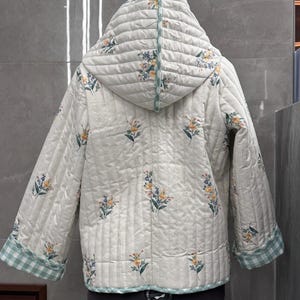 K&ouml;nnte beinhalten: Eine wei&szlig;e gesteppte Jacke mit Blumendruck und gr&uuml;nem und wei&szlig;em Karomuster. Die Jacke hat eine Kapuze und lange &Auml;rmel.