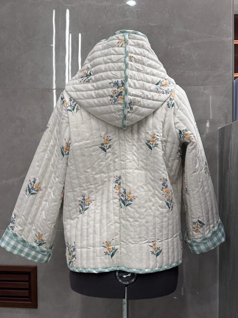 K&ouml;nnte beinhalten: Eine wei&szlig;e gesteppte Jacke mit Blumendruck und gr&uuml;nem und wei&szlig;em Karomuster. Die Jacke hat eine Kapuze und lange &Auml;rmel.