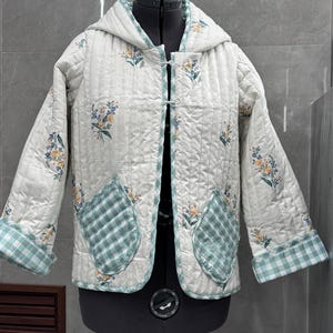 K&ouml;nnte beinhalten: Eine wei&szlig;e gesteppte Jacke mit Blumendruck und gr&uuml;nem Karomuster. Die Jacke hat eine Kapuze und zwei aufgesetzte Taschen.