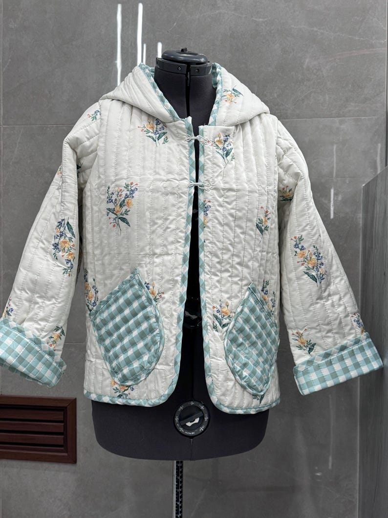 K&ouml;nnte beinhalten: Eine wei&szlig;e gesteppte Jacke mit Blumendruck und gr&uuml;nem Karomuster. Die Jacke hat eine Kapuze und zwei aufgesetzte Taschen.