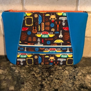 May include: A blue and brown zippered pouch with a Friends theme. The pouch is decorated with various images from the show, including the Central Perk logo, a coffee cup, a lobster, and a picture frame. The zipper is orange.