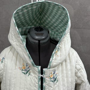 K&ouml;nnte beinhalten: Eine wei&szlig;e gesteppte Jacke mit einer gr&uuml;n-wei&szlig; karierten Kapuze und Blumendruck. Die Jacke hat einen Knopfverschluss und ist mit einem gr&uuml;n-wei&szlig; karierten Stoff gef&uuml;ttert.
