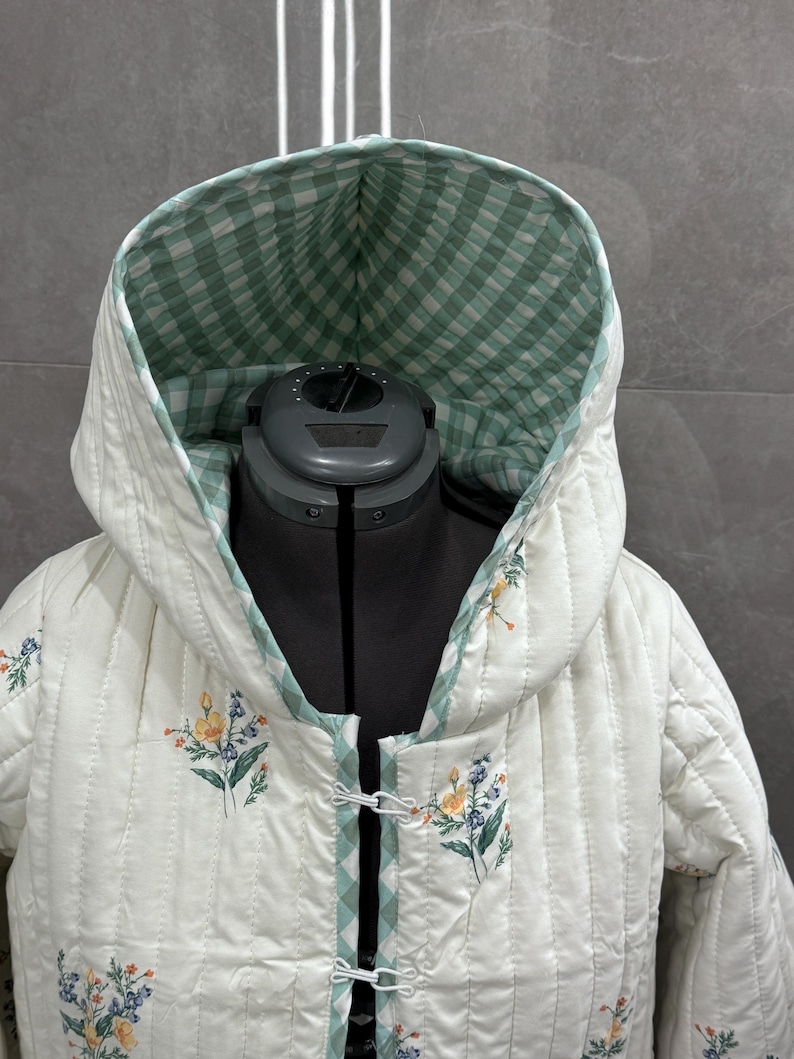 K&ouml;nnte beinhalten: Eine wei&szlig;e gesteppte Jacke mit einer gr&uuml;n-wei&szlig; karierten Kapuze und Blumendruck. Die Jacke hat einen Knopfverschluss und ist mit einem gr&uuml;n-wei&szlig; karierten Stoff gef&uuml;ttert.