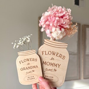 May include: Two wooden mason jar-shaped flower holders. One says "FLOWERS for GRANDMA" and the other says "FLOWERS for MOMMY". The Grandma jar is filled with white flowers, and the Mommy jar has pink and white dried flowers. The text "Picked by" is also on the jars.