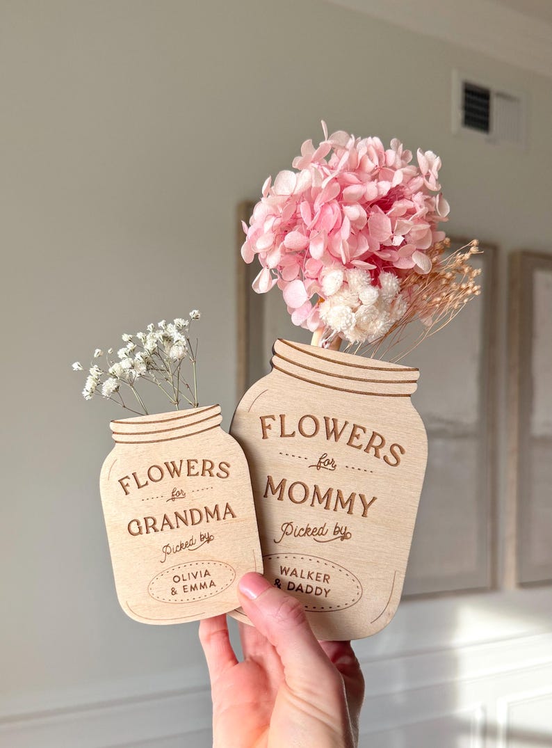 May include: Two wooden mason jar-shaped flower holders. One says "FLOWERS for GRANDMA" and the other says "FLOWERS for MOMMY". The Grandma jar is filled with white flowers, and the Mommy jar has pink and white dried flowers. The text "Picked by" is also on the jars.