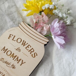 May include: A wooden mason jar-shaped sign with the words "FLOWERS for MOMMY" engraved on it. The sign is filled with a bouquet of yellow, pink, white, and purple flowers. The text "Picked by" and a name are also engraved on the sign.