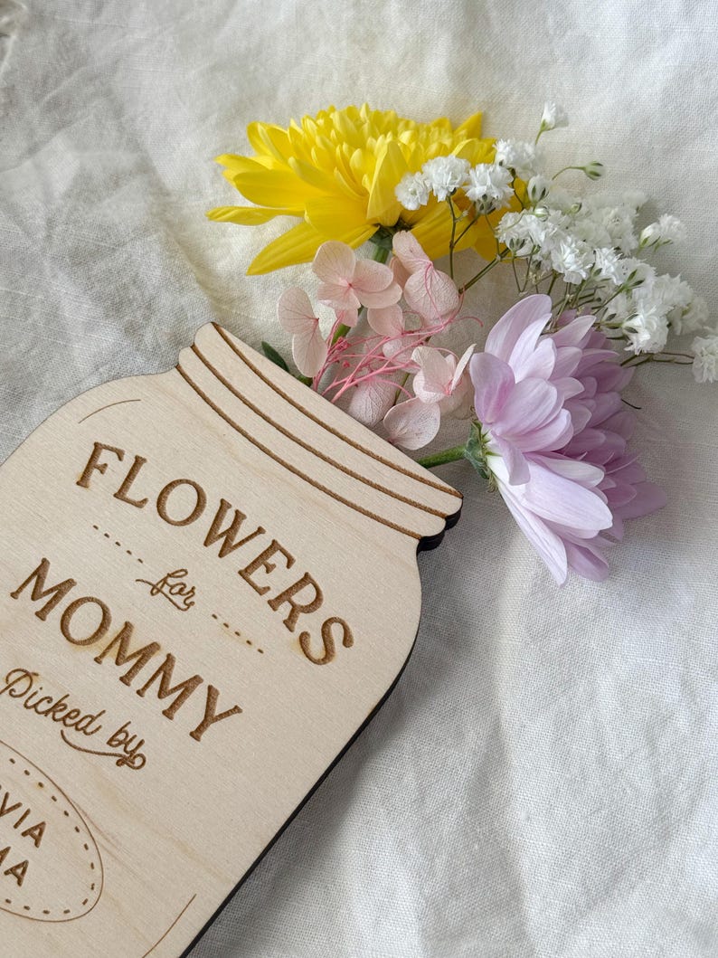May include: A wooden mason jar-shaped sign with the words "FLOWERS for MOMMY" engraved on it. The sign is filled with a bouquet of yellow, pink, white, and purple flowers. The text "Picked by" and a name are also engraved on the sign.