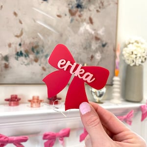 May include: A red bow-shaped decoration with the name "erika" in pink script. The bow is held in front of a white mantel decorated with pink bows. The background features a painting with neutral colors.