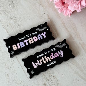 May include: Two black, wavy-edged signs with the text "boo! It's my BIRTHDAY" and "boo! It's my birthday witch" in pastel colors. The signs have glittery bat and star accents. Pink flowers are in the background.
