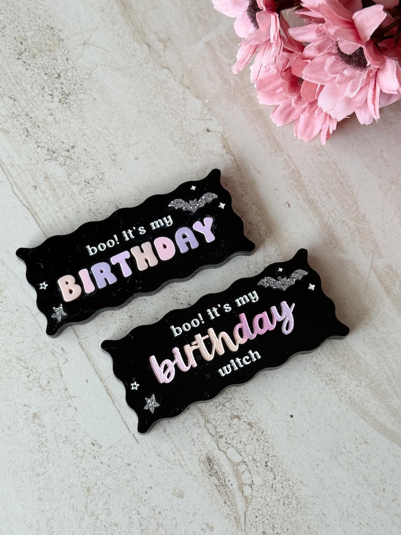 May include: Two black, wavy-edged signs with the text "boo! It's my BIRTHDAY" and "boo! It's my birthday witch" in pastel colors. The signs have glittery bat and star accents. Pink flowers are in the background.