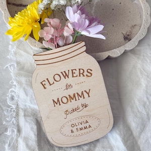 May include: A wooden mason jar-shaped sign with the words "FLOWERS for MOMMY Picked by OLIVIA & EMMA" engraved on it. The jar holds a bouquet of yellow, white, pink, and purple flowers. The sign is on a white surface.