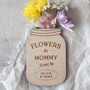 May include: A wooden mason jar-shaped sign with the words "FLOWERS for MOMMY Picked by OLIVIA & EMMA" engraved on it. The sign holds a small bouquet of yellow, white, pink, and purple flowers. The background is a white, textured fabric.