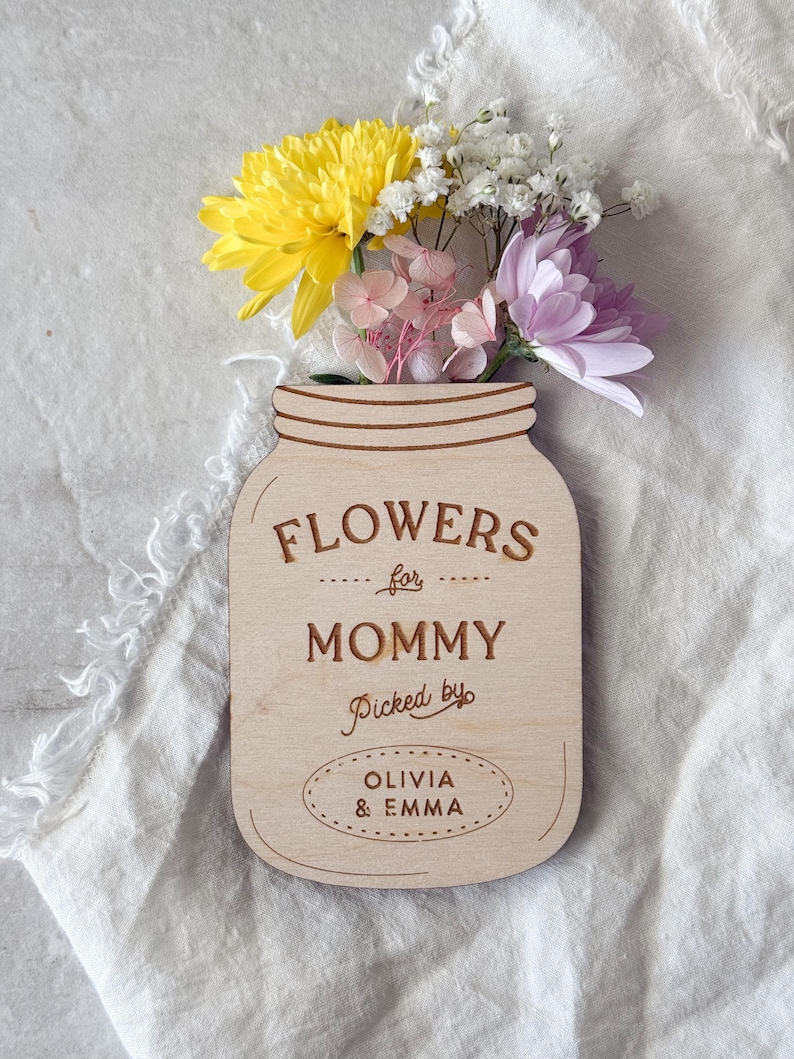 May include: A wooden mason jar-shaped sign with the words "FLOWERS for MOMMY Picked by OLIVIA & EMMA" engraved on it. The sign holds a small bouquet of yellow, white, pink, and purple flowers. The background is a white, textured fabric.
