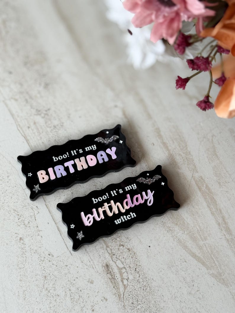 May include: Two black rectangular birthday-themed decorations. One reads "boo! It's my BIRTHDAY" in a gradient of pink and purple. The other says "boo! It's my birthday witch" in a similar gradient. Both have bat and star accents.