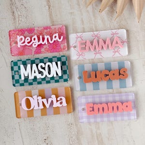 May include: Six rectangular acrylic name tags featuring various patterns and raised lettering. The tags display the names Regina, Emma, Mason, Lucas, Olivia, and Emma in different fonts, colors, and backgrounds including floral, bow, checkered, striped, and gingham prints.