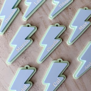 May include: A collection of light blue and green lightning bolt-shaped name tags. Each tag has a different name in white lettering, including "Thomas", "Leo", "Thiago", "Max", "Marco", "Charlie", "Nikhil", "Cal", and "And". The tags have a small hole at the top.