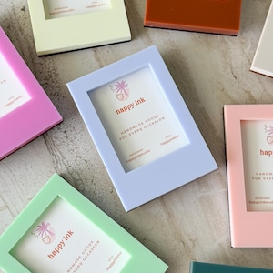 May include: A collection of colorful picture frames in various pastel shades, including pink, blue, green, and orange. Each frame has a white insert with the text "happy ink" and "HANDMADE GOODS FOR EVERY OCCASION".