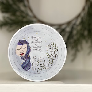 May include: A round sticker with a blue and purple illustration of a woman with closed eyes and long hair. The sticker has a white background with a gold outline and the text "You are the universe in ecstatic motion."