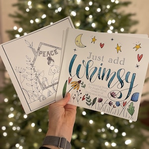May include: Two coloring books are held up in front of a blurred Christmas tree. One book has the word "PEACE" inside a geometric shape. The other book says "Just add Whimsy" with floral and celestial illustrations.