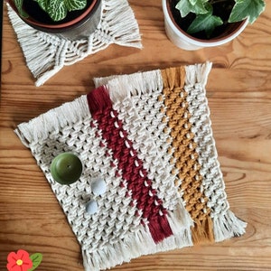 Könnte beinhalten: Handgefertigte Makramee-Untersetzer in Creme, Rot und Gold. Die Untersetzer haben ein gewebtes Design mit Fransen. Eine kleine grüne Kerze und zwei graue Steine liegen auf einem Untersetzer. Zwei Topfpflanzen im Hintergrund.