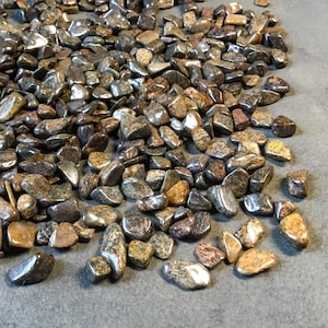 May include: A collection of polished tumbled stones in various shades of brown, black, and gold. The stones have a smooth, reflective surface and vary in size and shape. The image shows a close-up view of the stones, with some appearing to have a speckled or patterned texture.