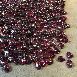 May include: A large pile of deep red, smooth, polished gemstones. The stones are scattered on a gray surface.