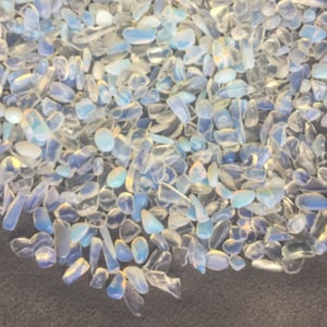 May include: A close-up of a pile of iridescent white and blue opal chips. The chips are various shapes and sizes, and they are scattered on a dark surface.