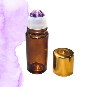 May include: A brown glass essential oil roller bottle with a purple amethyst rollerball and a gold cap. The bottle is open, with the cap next to it. The background has a purple watercolor design.