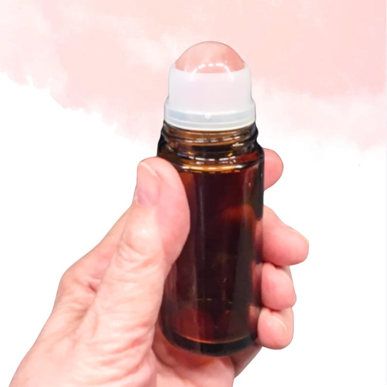 May include: A small, brown glass roll-on bottle with a white cap and a pink rollerball. The bottle is held in a hand against a soft pink and white background. The bottle appears to be filled with a liquid.