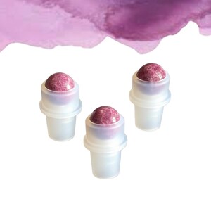 May include: Three clear roller bottles with a pink gemstone rollerball top. The background features a watercolour wash of pink and purple hues. The bottles are arranged in a triangular pattern.