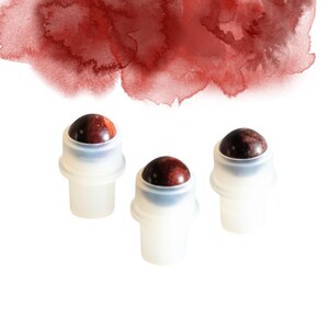 May include: Three roll-on bottles with dark red gemstone tops and white plastic bases. A deep red watercolour wash forms the background. The bottles are arranged in a triangular pattern.