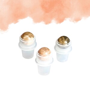 May include: Three clear glass roller bottles with gold and brown gemstone rollerballs. The bottles are arranged on a white surface with a peach-coloured watercolour wash in the background. The rollerballs have a marbled appearance.