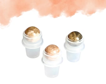 Könnte beinhalten: Drei transparente Glas-Rollerflaschen mit gold- und braunfarbenen Edelstein-Rollerkugeln. Die Flaschen sind auf einer weißen Oberfläche mit einem pfirsichfarbenen Aquarell-Waschgang im Hintergrund angeordnet. Die Rollerkugeln haben ein marmoriertes Aussehen.