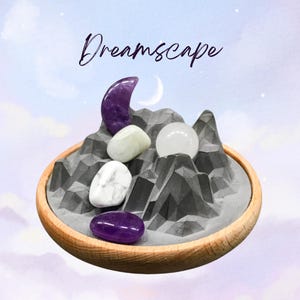 May include: A wooden-rimmed tray holds a gray mountain-shaped sculpture with various gemstones. The gemstones include a purple amethyst crescent, white and green stones, and a clear sphere. The word "Dreamscape" is written in cursive above.