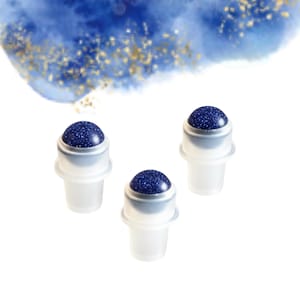 May include: Three facial roller bottles with a dark blue, speckled stone top. The roller bottles have a clear, frosted plastic base and a silver metal rim. The background features a blue watercolor wash with gold flecks.