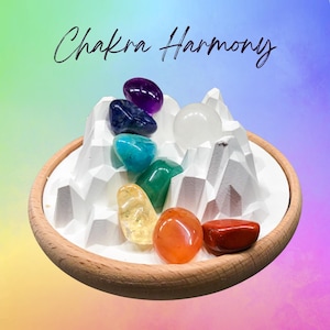 May include: A wooden tray with a white, mountain-shaped structure holds seven polished chakra stones in various colors. The stones are arranged in a semi-circle, with the text "Chakra Harmony" in cursive script above.