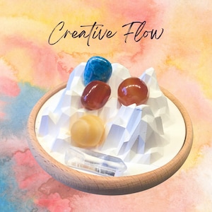 以下が含まれることがあります： 木製の縁が付いたトレイには、さまざまな色の宝石が置かれた白い幾何学的な山のデザインがあります。 宝石には、青、赤、黄色の石と透明なクリスタルが含まれています。 テキスト「Creative Flow」は筆記体で書かれています。