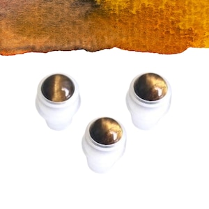 May include: Three small, white, oval-shaped objects with a polished, round, brown and gold stone set in the center. The background is white with a watercolour wash of orange and brown.
