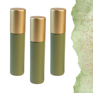 Puede incluir: Tres botellas roll-on de color verde oliva con tapas doradas. Las botellas son cilíndricas y se alzan sobre un fondo blanco con un diseño de acuarela verde en la esquina superior derecha.