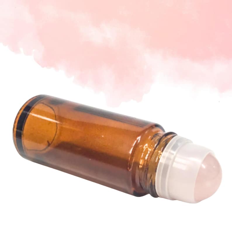 May include: A brown glass roller bottle with a white cap and a pink rollerball. The bottle is lying on its side, and the background is a soft pink and white.