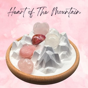 Pode incluir: Uma bandeja de madeira apresenta uma exibição branca em forma de montanha com várias pedras preciosas rosa e vermelhas. As pedras preciosas incluem um quartzo rosa em forma de coração, um quartzo claro redondo e outras pedras polidas. O texto "Heart of The Mountain" está escrito em itálico.