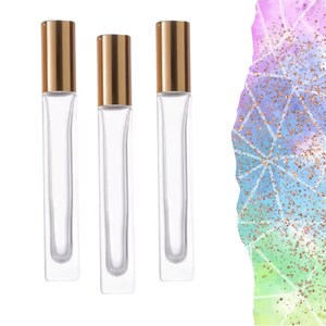 May include: Three clear glass perfume or essential oil roller bottles with gold-coloured caps. The bottles are rectangular and empty. The background features a colourful abstract design with geometric shapes and glitter.