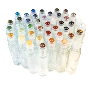 May include: A collection of clear glass essential oil roller bottles with various gemstone tops. The gemstones include blue, green, red, and brown hues. The bottles are arranged closely together against a white background.