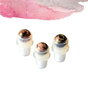 May include: Three cosmetic roller bottles with a gemstone rollerball top. The bottles are white and clear, and the gemstone is pink and brown. The background has a pink and grey watercolour wash.