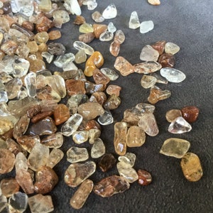 May include: A collection of small, translucent, and opaque, light brown, and clear quartz crystals. The crystals are scattered on a dark gray surface.
