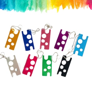Op de afbeelding: Een verzameling kleurrijke, rechthoekige sleutelhangers in verschillende kleuren, waaronder goud, blauw, roze, paars, zilver, rood, groen, zwart en lichtblauw. Elke sleutelhanger heeft een uniek ontwerp met drie cirkelvormige uitsparingen en een kleine ketting.