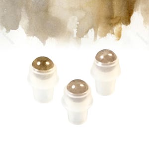 May include: Three small, clear rollerball applicators with metallic, rounded tops. The applicators are set against a white background with a watercolour wash of brown and tan hues in the upper portion of the image.