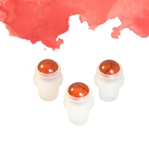 May include: Three roll-on bottles with orange-red gemstone tops. The bottles have a frosted white base and are set against a white background with a red watercolor wash in the upper portion of the image.
